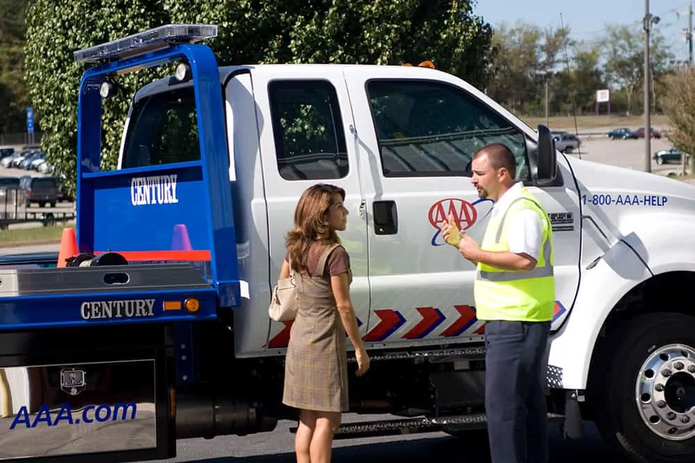 A photo of a AAA service worker speaking with a woman who may be dealing with car troubles. The pair are pictured conversing on the side of a road in front of the AAA tow truck. 