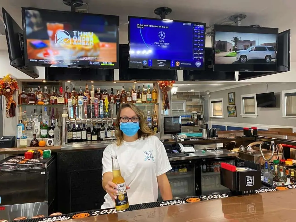 A woman wearing a facemask and holding a corona beer out towards the camera, standing behind a bar with three televisions above hear head and a wide variety of alcoholic beverages shown behind her. 